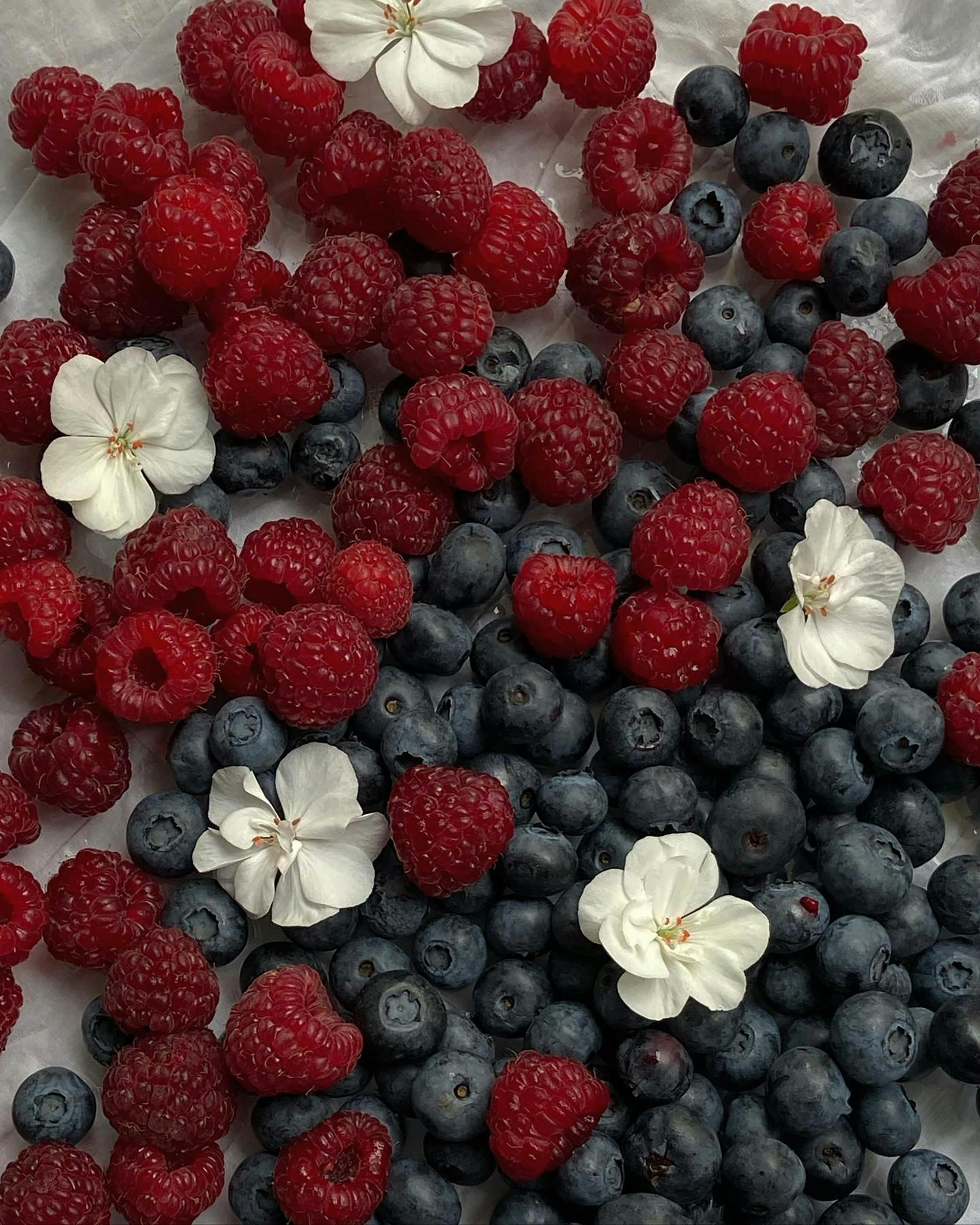 Ein lebendiges, flach arrangiertes Bild von frischen Blaubeeren, Himbeeren und weißen Blüten auf hellem Hintergrund.